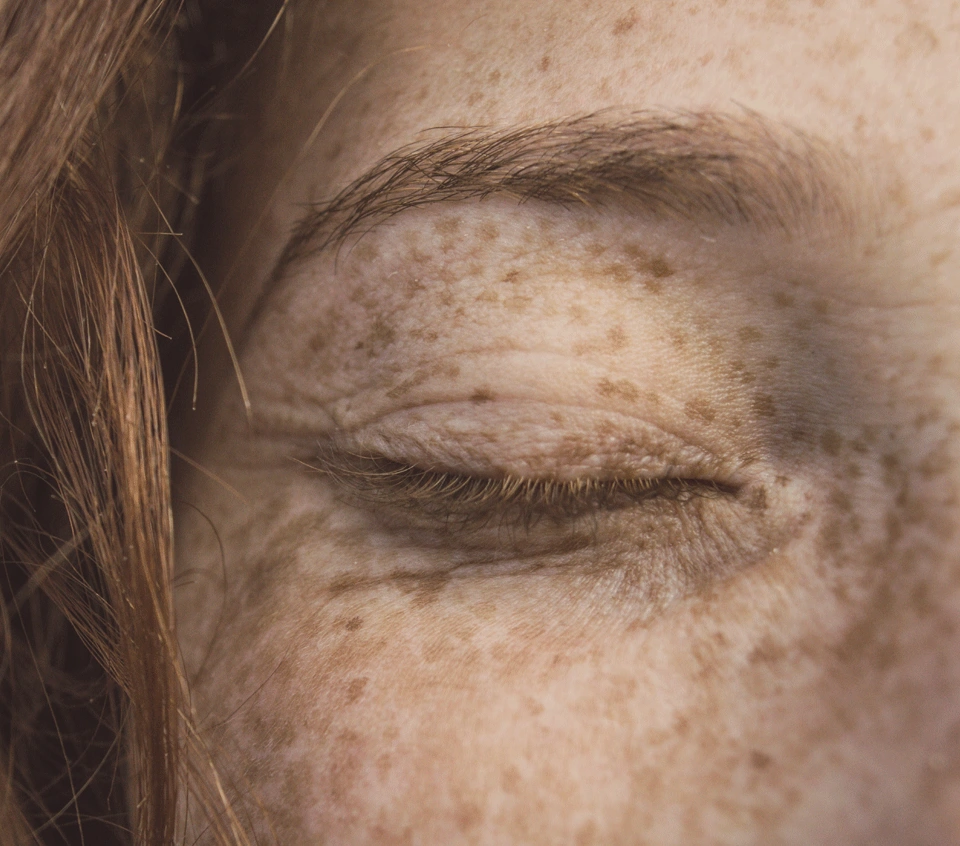 Nahaufnahme einer Frau mit rotem Haar, Sommersprossen und geschlossenen Augen: Hautstruktur, Augenlid, Brauen und lange Wimpern. Authentische Darstellung der Gesichtsmerkmale.