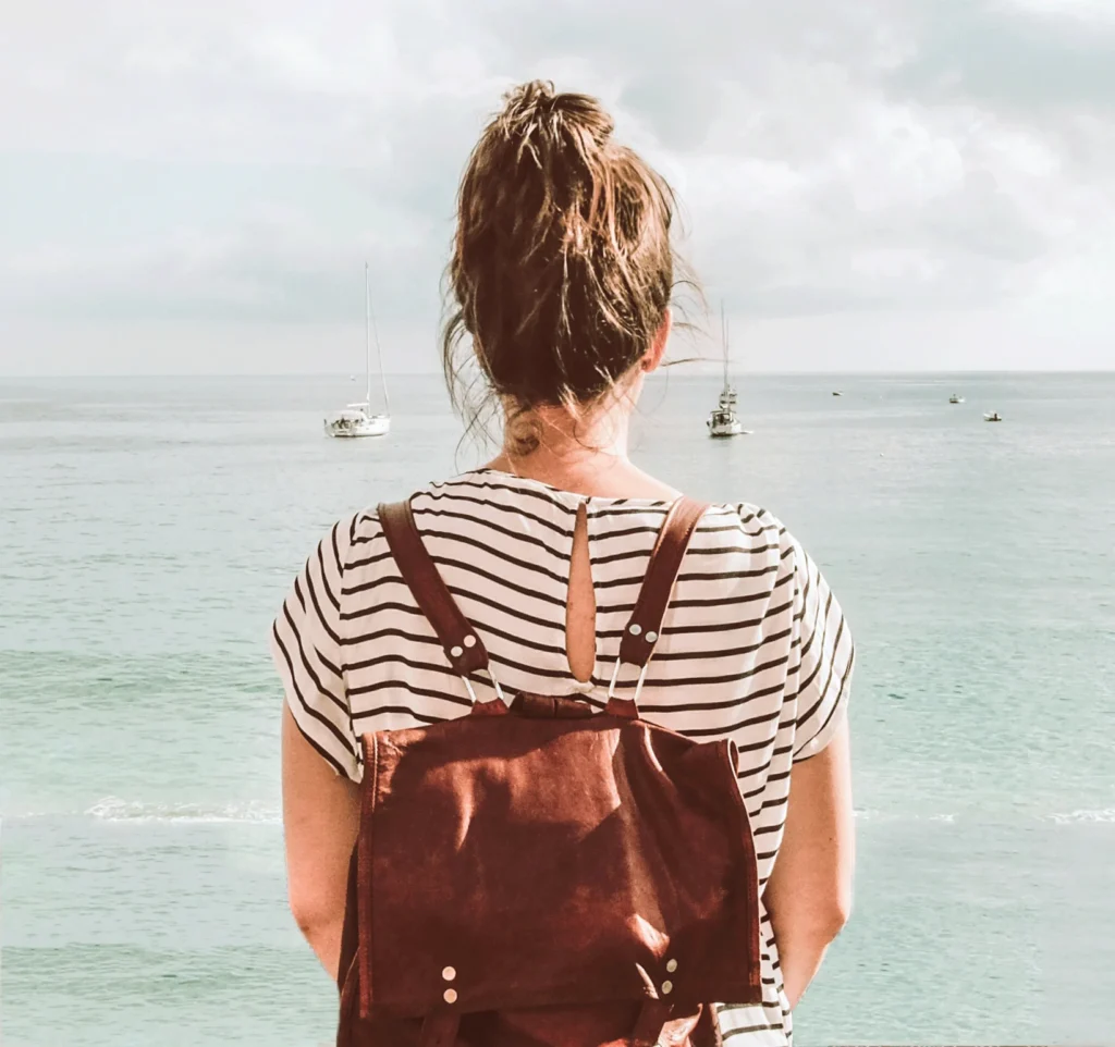 Rückansicht einer Frau mit Pferdeschwanz und braunem Rucksack schaut aufs Meer.